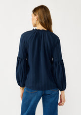 [Color: Midnight] A back facing image of a blonde model wearing a blue long sleeve peasant blouse with a subtle checkered pattern. Featuring long bishop sleeves, ruffled neckline, tie detail, and button front. 