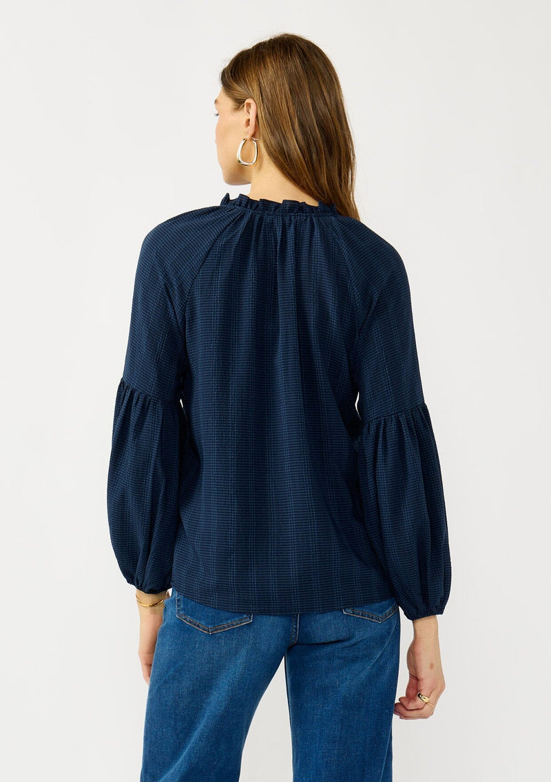 [Color: Midnight] A back facing image of a blonde model wearing a blue long sleeve peasant blouse with a subtle checkered pattern. Featuring long bishop sleeves, ruffled neckline, tie detail, and button front. 