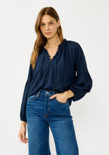 [Color: Midnight] A front facing image of a blonde model wearing a blue long sleeve peasant blouse with a subtle checkered pattern. Featuring long bishop sleeves, ruffled neckline, tie detail, and button front. 