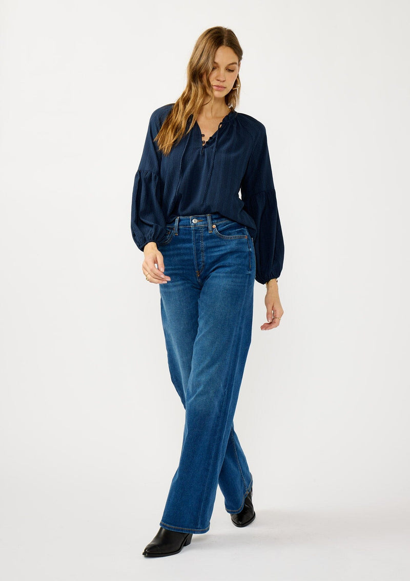 [Color: Midnight] A front facing image of a blonde model wearing a blue long sleeve peasant blouse with a subtle checkered pattern. Featuring long bishop sleeves, ruffled neckline, tie detail, and button front. 