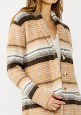 [Color: Camel/Brown] A detail image of a blonde model wearing a brown and tan striped mid length coatigan with a shawl collar, button front, and side patch pockets. Effortlessly cozy for the fall and winter season. 