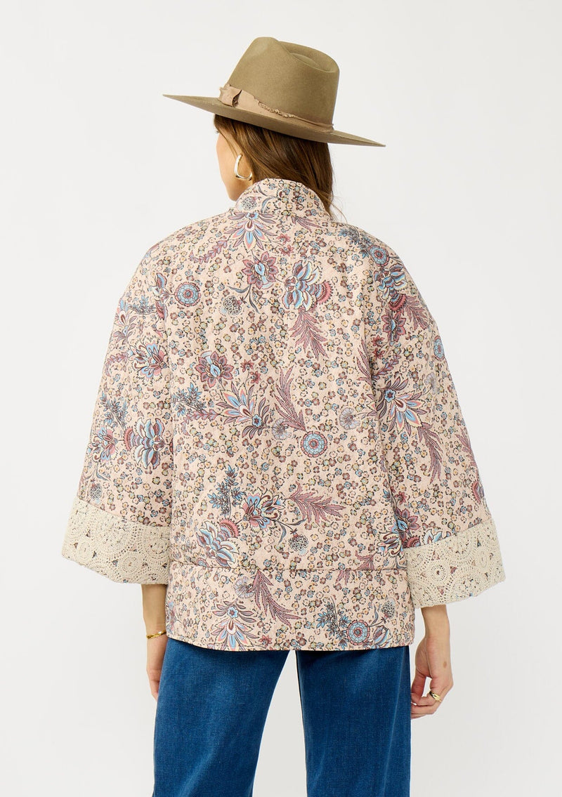 [Color: Mauve/Natural] A back facing image of a blonde model wearing a mauve pink floral bohemian jacket with an oversized fit, long sleeves, crochet trim, and side pockets. 