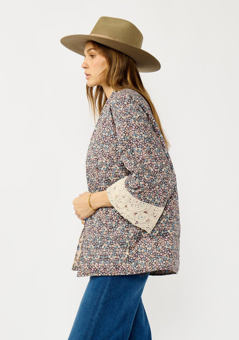 [Color: Mauve/Natural] A side facing image of a blonde model wearing a reversible mauve pink floral bohemian jacket with an oversized fit, long sleeves, crochet trim, and side pockets. 