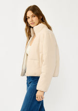 [Color: Natural] A side facing image of a blonde model wearing a light beige soft knit puffer jacket with a zipper front, high collar, and side pockets. A classic fall jacket for cold weather. 