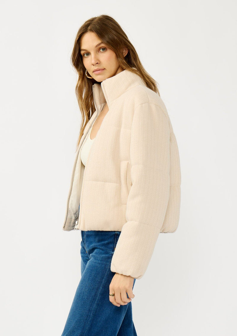 [Color: Natural] A side facing image of a blonde model wearing a light beige soft knit puffer jacket with a zipper front, high collar, and side pockets. A classic fall jacket for cold weather. 