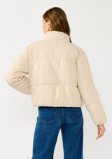 [Color: Natural] A back facing image of a blonde model wearing a light beige soft knit puffer jacket with a zipper front, high collar, and side pockets. A classic fall jacket for cold weather. 