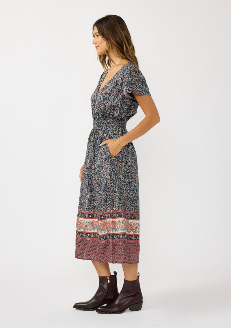 [Color: Navy/Rose] A side facing image of a brunette model wearing a navy floral border print midi dress with a classic v-neckline, short sleeves, a smocked waist, and side pockets. A fall bohemian dress perfect for any occasion. 