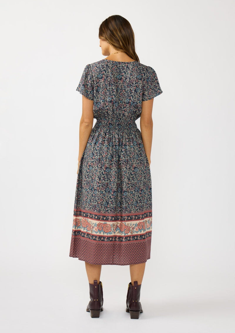 [Color: Navy/Rose] A back facing image of a brunette model wearing a navy floral border print midi dress with a classic v-neckline, short sleeves, a smocked waist, and side pockets. A fall bohemian dress perfect for any occasion. 