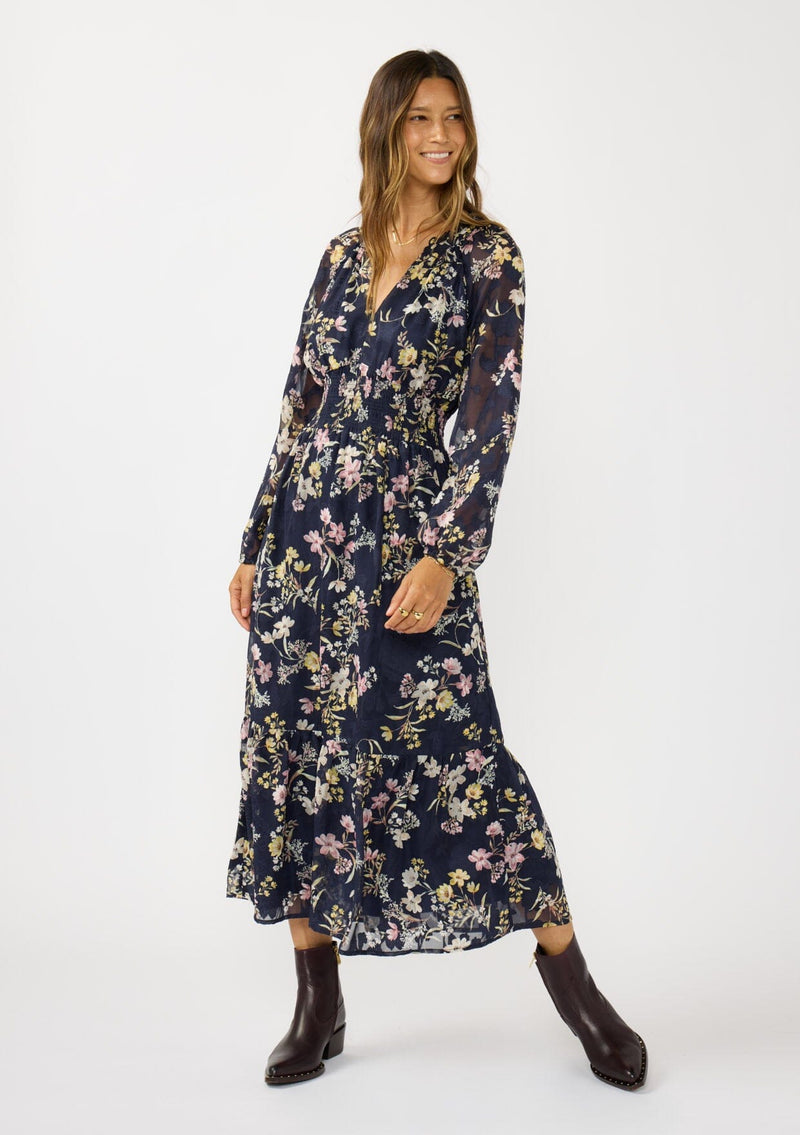 [Color: Navy/Yellow] A front facing image of a brunette model wearing a navy floral maxi dress with long sleeves, a smocked waist, and a split v-neckline. A classic floral maxi for weddings and special occasions. 