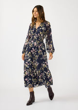 [Color: Navy/Yellow] A front facing image of a brunette model wearing a navy floral maxi dress with long sleeves, a smocked waist, and a split v-neckline. A classic floral maxi for weddings and special occasions. 