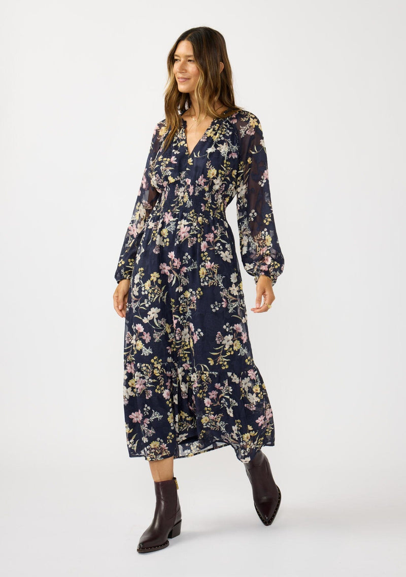 [Color: Navy/Yellow] A front facing image of a brunette model wearing a navy floral maxi dress with long sleeves, a smocked waist, and a split v-neckline. A classic floral maxi for weddings and special occasions. 