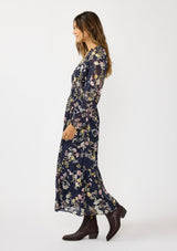 [Color: Navy/Yellow] A side facing image of a brunette model wearing a navy floral maxi dress with long sleeves, a smocked waist, and a split v-neckline. A classic floral maxi for weddings and special occasions. 