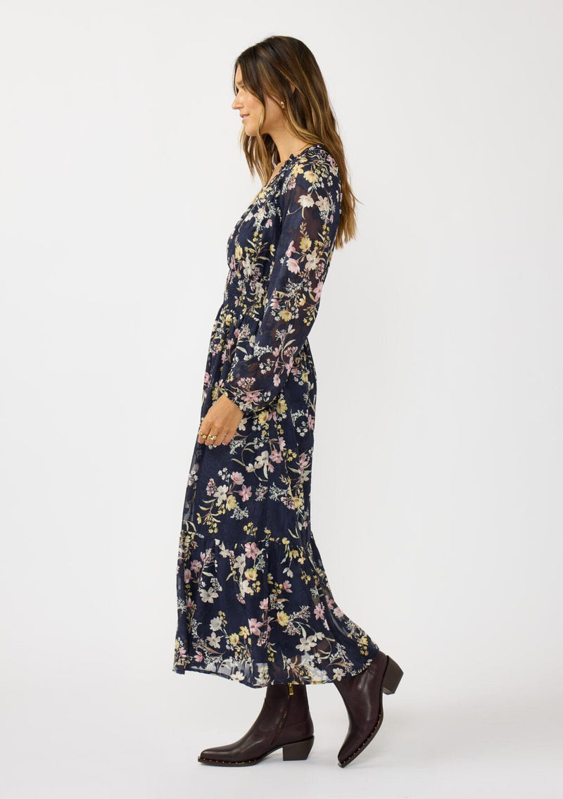 [Color: Navy/Yellow] A side facing image of a brunette model wearing a navy floral maxi dress with long sleeves, a smocked waist, and a split v-neckline. A classic floral maxi for weddings and special occasions. 