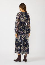 [Color: Navy/Yellow] A back facing image of a brunette model wearing a navy floral maxi dress with long sleeves, a smocked waist, and a split v-neckline. A classic floral maxi for weddings and special occasions. 