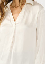 [Color: Eggshell] A detail image of a brunette model wearing an ivory silky poly satin blouse with a split v-neckline, collar, and long sleeves with button cuffs. 