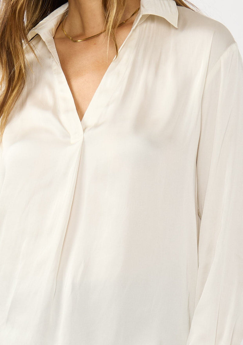 [Color: Eggshell] A detail image of a brunette model wearing an ivory silky poly satin blouse with a split v-neckline, collar, and long sleeves with button cuffs. 