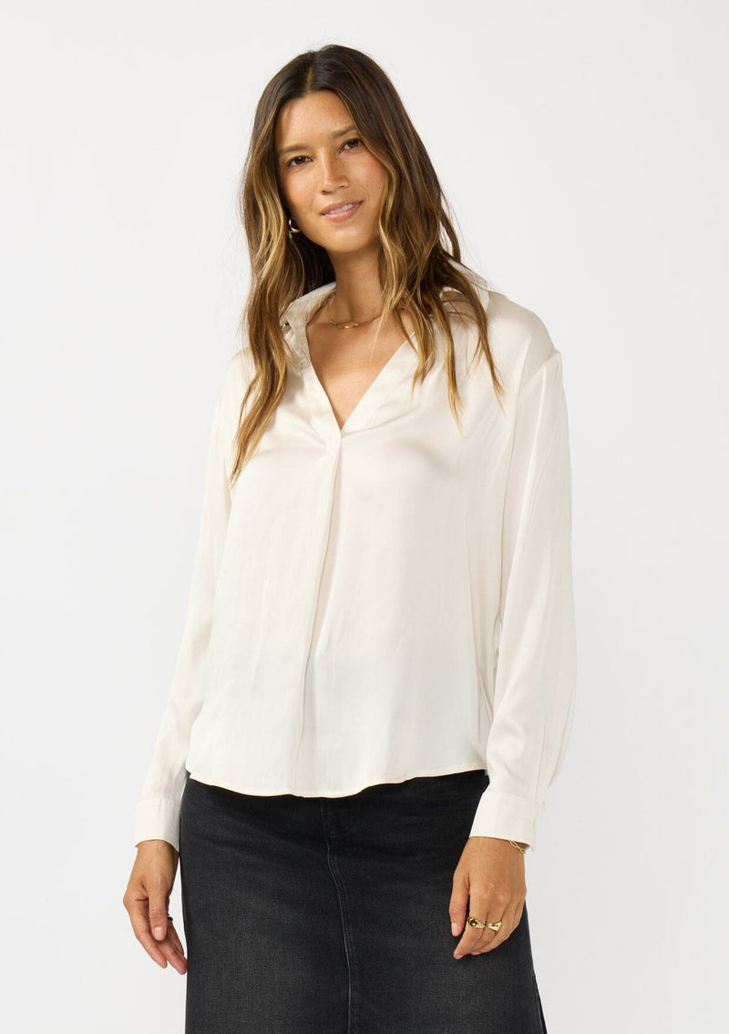 [Color: Eggshell] A front facing image of a brunette model wearing an ivory silky poly satin blouse with a split v-neckline, collar, and long sleeves with button cuffs. 
