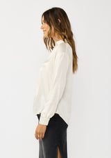 [Color: Eggshell] A side facing image of a brunette model wearing an ivory silky poly satin blouse with a split v-neckline, collar, and long sleeves with button cuffs. 