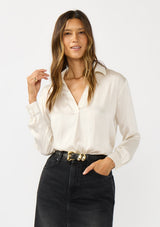 [Color: Eggshell] A front facing image of a brunette model wearing an ivory silky poly satin blouse with a split v-neckline, collar, and long sleeves with button cuffs. 