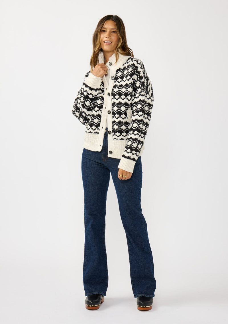 [Color: Cream/Black] A front facing image of a brunette model wearing a black and cream button front knit sweater with a high neckline and side pockets. A cold weather layering piece for the fall and winter season. 