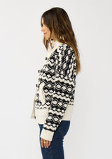 [Color: Cream/Black] A side facing image of a brunette model wearing a black and cream button front knit sweater with a high neckline and side pockets. A cold weather layering piece for the fall and winter season. 