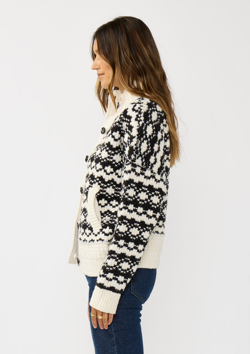 [Color: Cream/Black] A side facing image of a brunette model wearing a black and cream button front knit sweater with a high neckline and side pockets. A cold weather layering piece for the fall and winter season. 