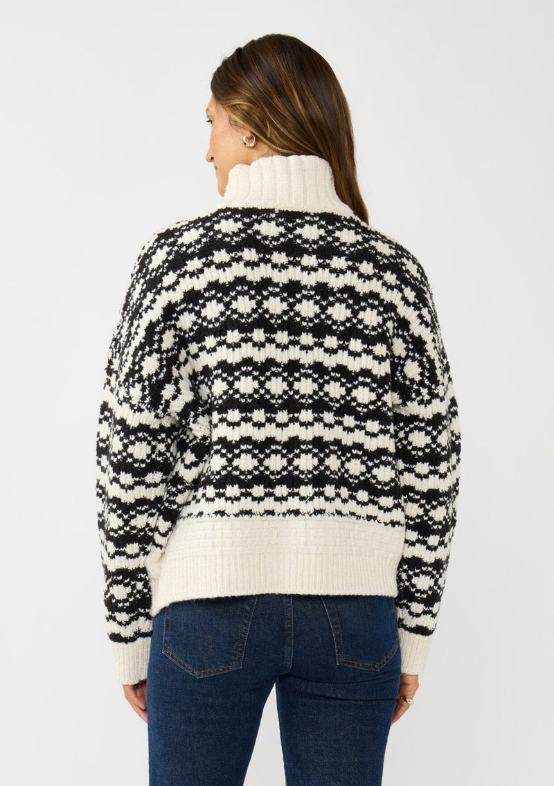 [Color: Cream/Black] A back facing image of a brunette model wearing a black and cream button front knit sweater with a high neckline and side pockets. A cold weather layering piece for the fall and winter season. 