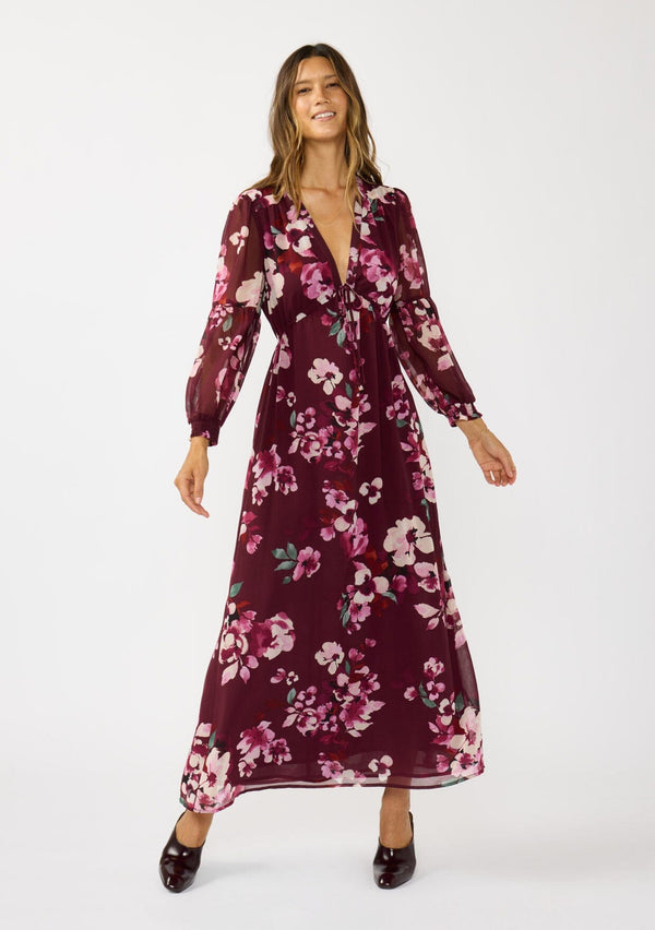 [Color: Wine/Dusty Rose] A front facing image of a brunette model wearing a burgundy floral maxi dress with an empire waist with drawstring, long sleeves, and a deep v-neckline. An elegant dress for the holidays or other special occasions. 