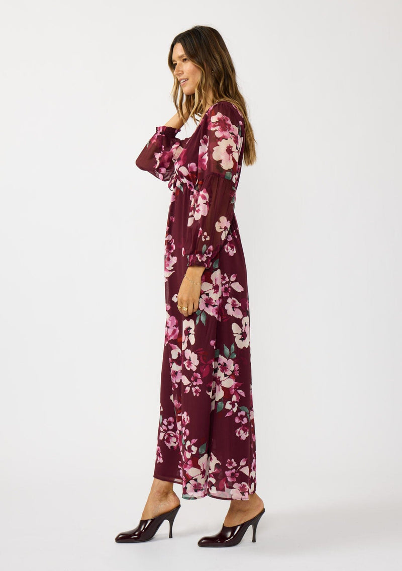 [Color: Wine/Dusty Rose] A side facing image of a brunette model wearing a burgundy floral maxi dress with an empire waist with drawstring, long sleeves, and a deep v-neckline. An elegant dress for the holidays or other special occasions. 
