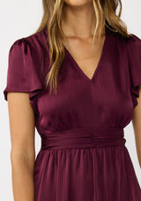 [Color: Merlot] A  detail image of a brunette model wearing a burgundy holiday midi dress with a short flutter sleeves, a defined waistline with gathered fabric, and a ruffled tiered skirt. Elegant and formal for date nights, holiday parties, and other special occasions. 