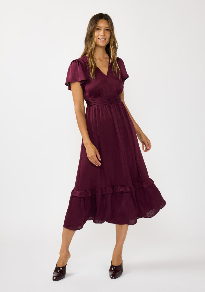 [Color: Merlot] A front facing image of a brunette model wearing a burgundy holiday midi dress with a short flutter sleeves, a defined waistline with gathered fabric, and a ruffled tiered skirt. Elegant and formal for date nights, holiday parties, and other special occasions. 