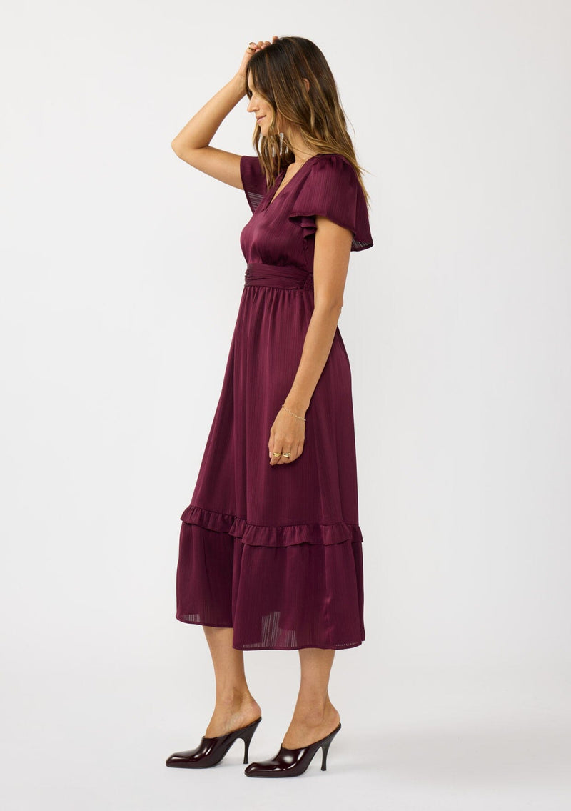 [Color: Merlot] A  side facing image of a brunette model wearing a burgundy holiday midi dress with a short flutter sleeves, a defined waistline with gathered fabric, and a ruffled tiered skirt. Elegant and formal for date nights, holiday parties, and other special occasions. 