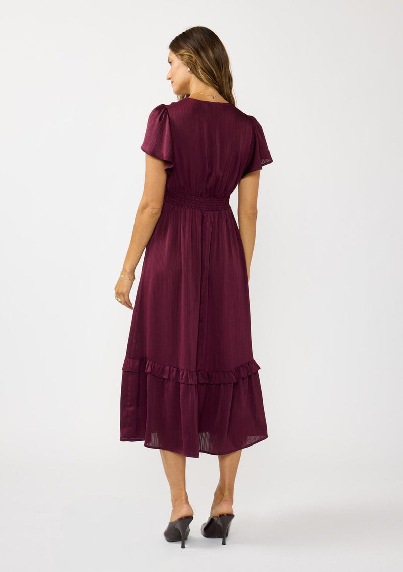 [Color: Merlot] A back facing image of a brunette model wearing a burgundy holiday midi dress with a short flutter sleeves, a defined waistline with gathered fabric, and a ruffled tiered skirt. Elegant and formal for date nights, holiday parties, and other special occasions. 