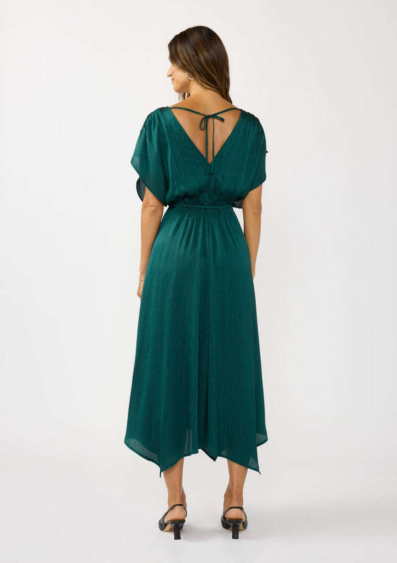 [Color: Emerald] A front facing image of a brunette model wearing a formal holiday party dress with sparkly metallic stripe details. Featuring a surplice v-neckline, short dolman sleeves, an elastic waist, open back with tie detail,  and an asymmetrical hemline. 