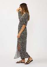 [Color: Black/Natural] A side facing image of a brunette model wearing a black and white abstract maxi dress with a deep v-neckline, half sleeves, empire waist, and a sexy side slit.