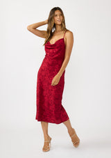[Color: Red] A front facing image of a brunette model wearing a holiday floral jacquard midi dress with a cowl neckline, adjustable straps, and a cross back. Elegant and timeless for holiday parties, weddings, and other special occasions. 