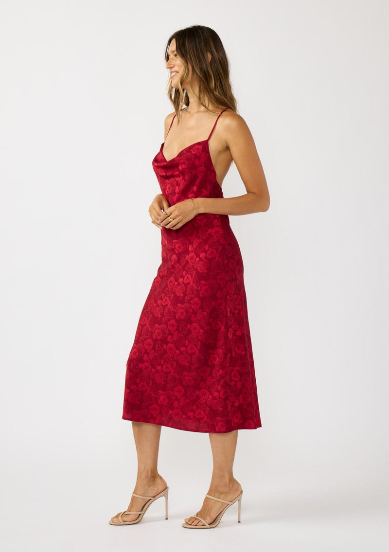 [Color: Red] A side facing image of a brunette model wearing a holiday floral jacquard midi dress with a cowl neckline, adjustable straps, and a cross back. Elegant and timeless for holiday parties, weddings, and other special occasions. 