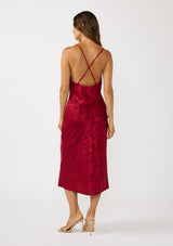 [Color: Red] A back facing image of a brunette model wearing a holiday floral jacquard midi dress with a cowl neckline, adjustable straps, and a cross back. Elegant and timeless for holiday parties, weddings, and other special occasions. 