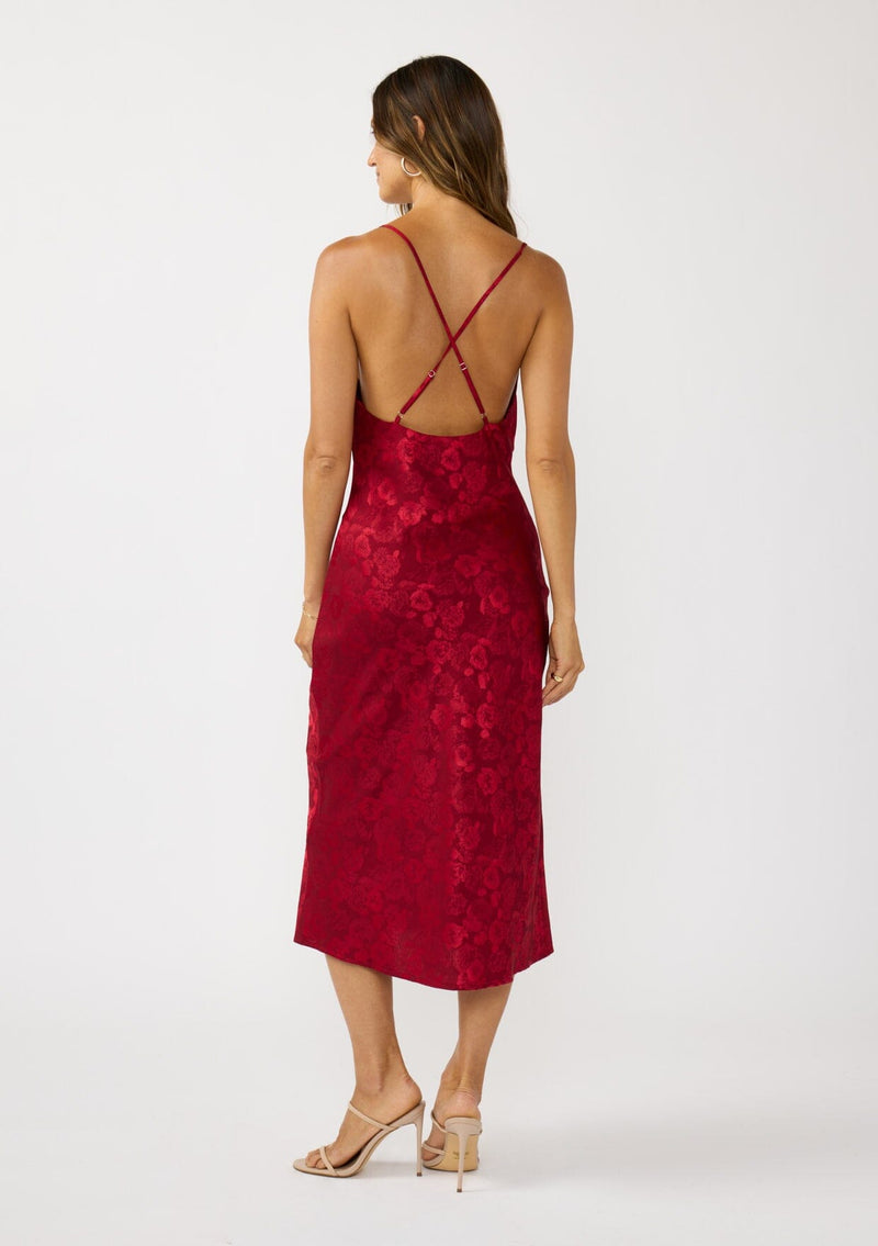 [Color: Red] A back facing image of a brunette model wearing a holiday floral jacquard midi dress with a cowl neckline, adjustable straps, and a cross back. Elegant and timeless for holiday parties, weddings, and other special occasions. 