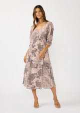 [Color: Natural/Brown] A front facing image of a brunette model wearing a nude brown floral midi dress with a v-neckline, 3/4 length sleeves, and a flattering empire waist. 