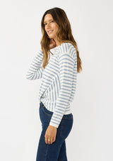 [Color: Ivory/Dusty Blue] A side facing image of a brunette model wearing a classic ivory and light blue striped knit pullover sweater. With long sleeves, a drop shoulder, a crew neckline, and a twist front detail at the waist. 