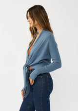 [Color: Heather Ocean] A photo of a Lovestitch model wearing a chic blue waffle knit wrap front sweater with long sleeves, a v neckline, and a button closure at the back. The long ties can be styled in multiple ways.