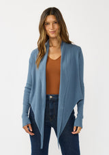 [Color: Heather Ocean] A photo of a Lovestitch model wearing a chic blue waffle knit wrap front sweater with long sleeves, a v neckline, and a button closure at the back. The long ties can be styled in multiple ways.