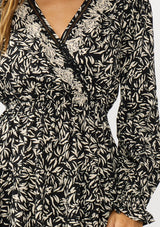 [Color: Black/Taupe] A detail image of a brunette model wearing an abstract ivory and black mini dress with embroidered top. Featuring long sleeves, elastic ruffled cuffs, a surplice v-neckline, elastic waist, and a ruffle trimmed tiered hem. 