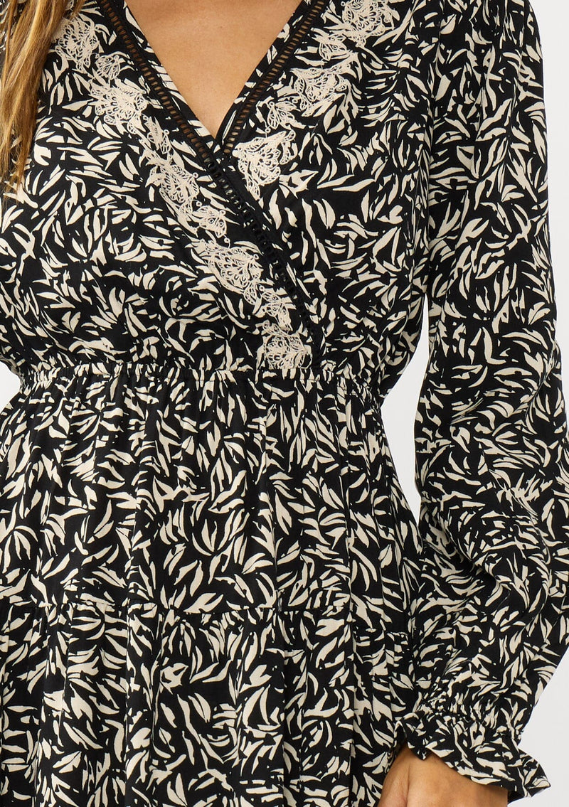 [Color: Black/Taupe] A detail image of a brunette model wearing an abstract ivory and black mini dress with embroidered top. Featuring long sleeves, elastic ruffled cuffs, a surplice v-neckline, elastic waist, and a ruffle trimmed tiered hem. 