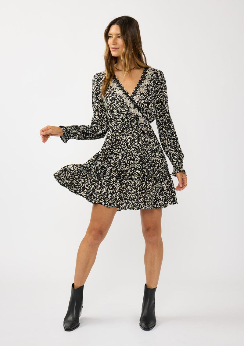 [Color: Black/Taupe] A front facing image of a brunette model wearing an abstract ivory and black mini dress with embroidered top. Featuring long sleeves, elastic ruffled cuffs, a surplice v-neckline, elastic waist, and a ruffle trimmed tiered hem. 