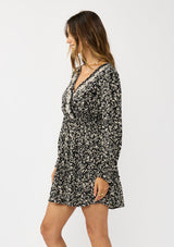 [Color: Black/Taupe] A side facing image of a brunette model wearing an abstract ivory and black mini dress with embroidered top. Featuring long sleeves, elastic ruffled cuffs, a surplice v-neckline, elastic waist, and a ruffle trimmed tiered hem. 