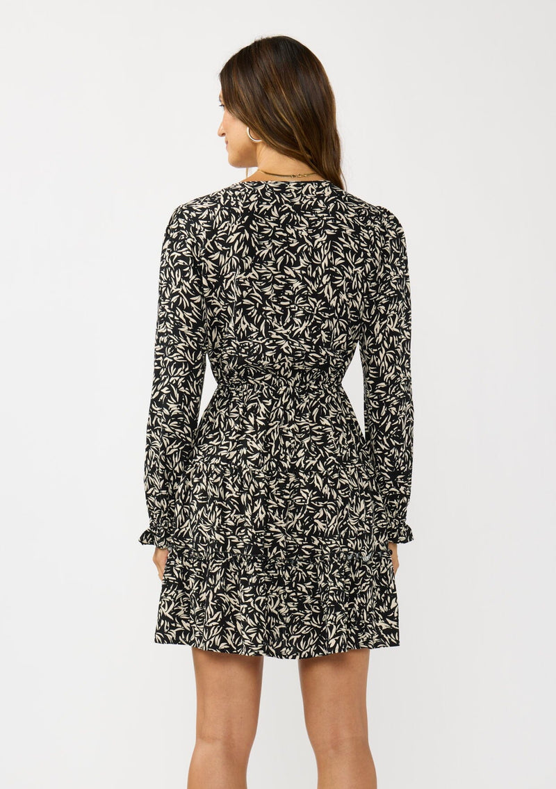 [Color: Black/Taupe] A back facing image of a brunette model wearing an abstract ivory and black mini dress with embroidered top. Featuring long sleeves, elastic ruffled cuffs, a surplice v-neckline, elastic waist, and a ruffle trimmed tiered hem. 