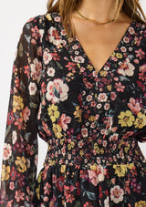 [Color: Black/Rust] A detail image of a brunette model wearing a black floral mini dress with a smocked waist, v-neckline, and long sleeves with smocked cuffs. A feminine and flirty mini dress for weddings, holidays, or other special occasions. 
