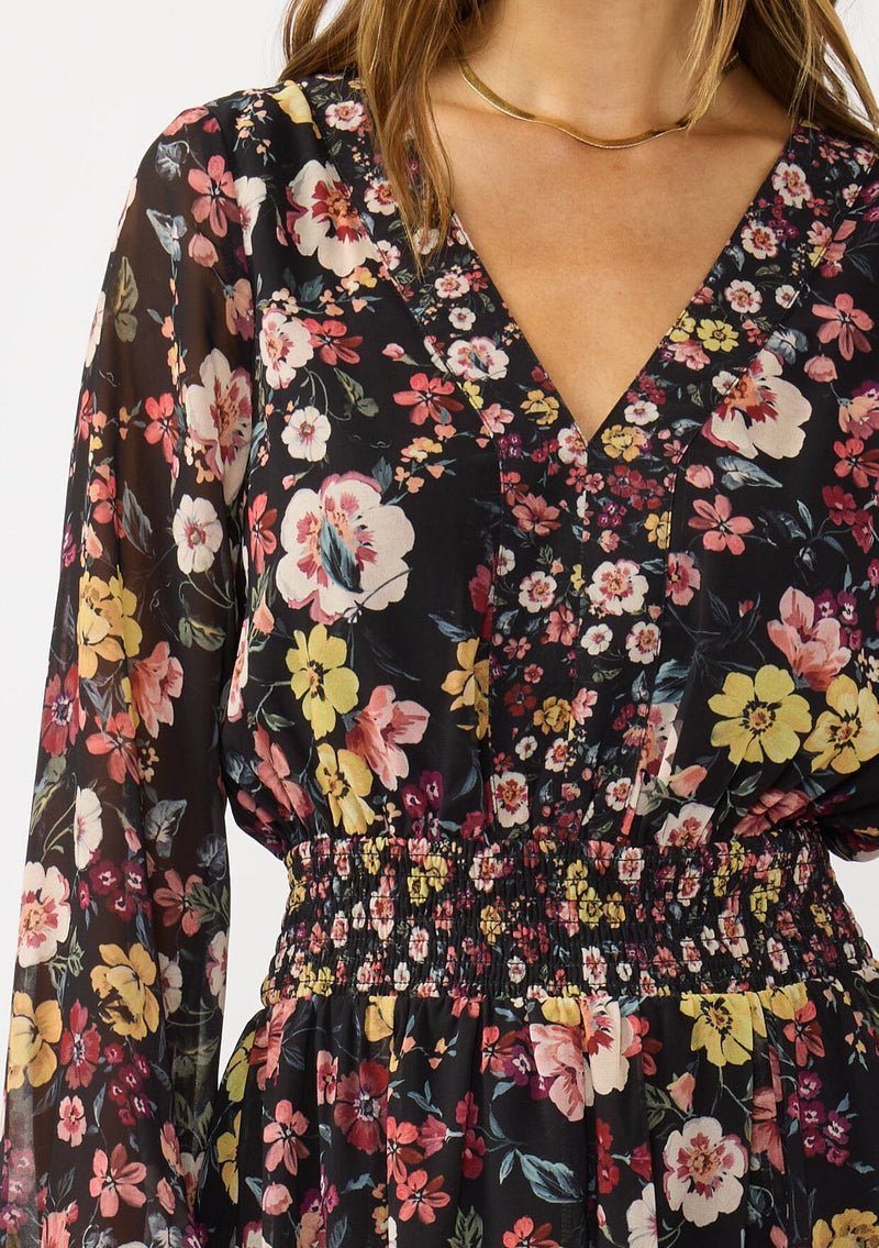 [Color: Black/Rust] A detail image of a brunette model wearing a black floral mini dress with a smocked waist, v-neckline, and long sleeves with smocked cuffs. A feminine and flirty mini dress for weddings, holidays, or other special occasions. 